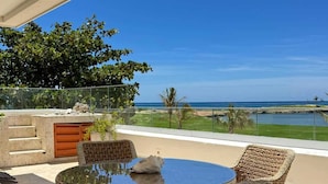 Eine überdachte Terrasse mit Korbmöbeln, einem Glastisch und Blick auf das Meer.