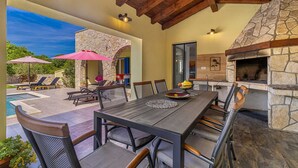 An outdoor patio with a stone fireplace, a dining table, and lounge chairs. - Holiday house MARLY with private pool (Kras)