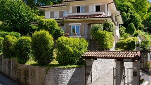 A two-story house with a stone wall and a tiled roof, surrounded by well-maintained bushes and trees. - LAKE Major Gem Gold (Stresa)
