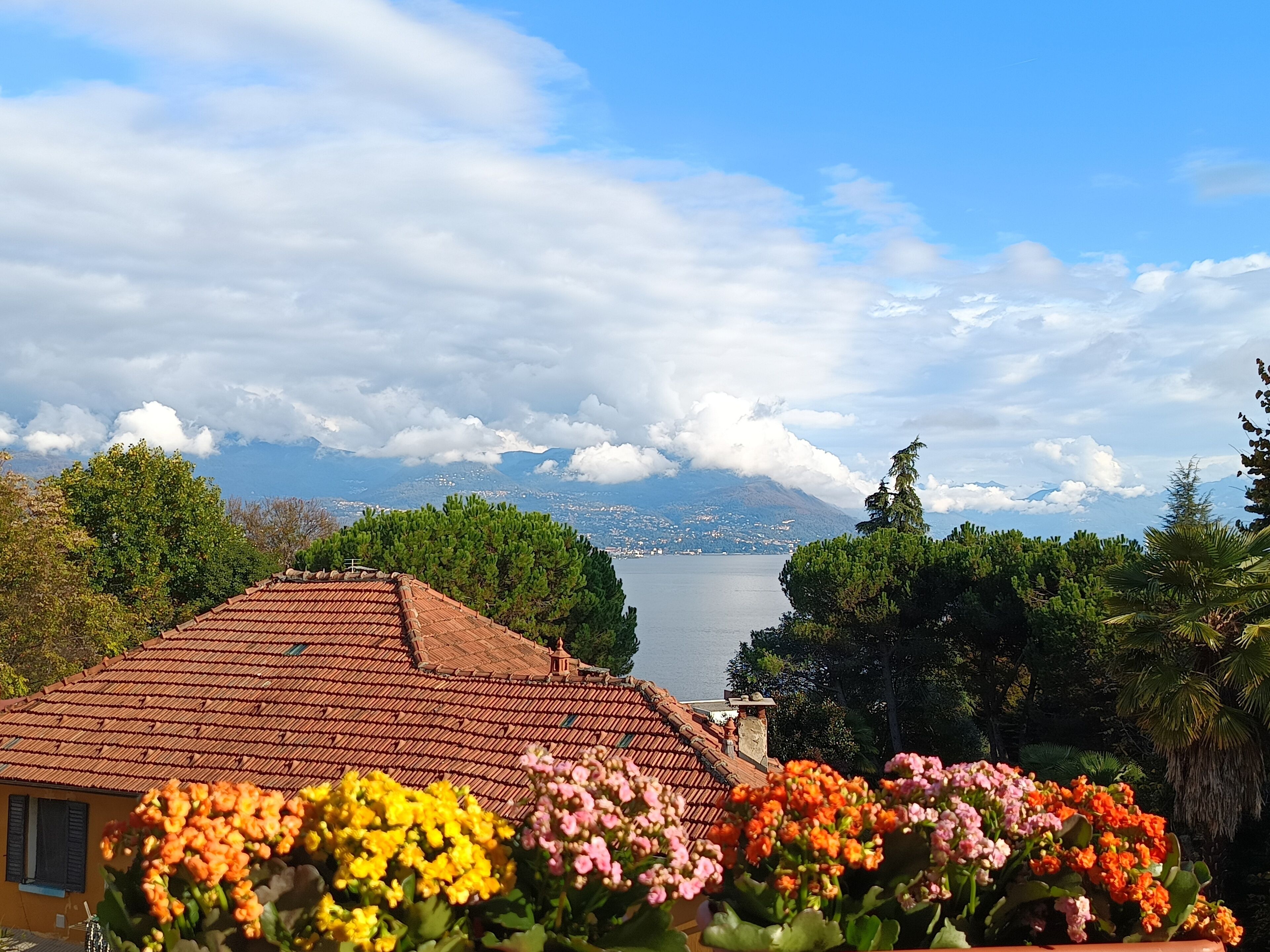 A house with a tiled roof, surrounded by greenery and flowers, overlooking a lake and mountains.