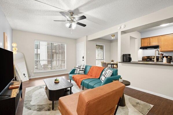 A modern living room with a sofa, coffee table, and a TV. There is a kitchen area with wooden cabinets and a countertop.