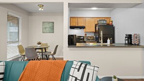 A modern kitchen with wooden cabinets, a black refrigerator, and a microwave. There is a dining area with a table and chairs, and a living room with a teal sofa and an orange throw blanket. - Austin Escape • Pool • 5 mins to Hyde Park UT (Austin)