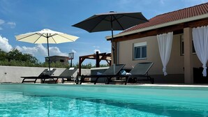 Ein Schwimmbecken mit klarem Wasser, umgeben von Liegestühlen und Sonnenschirmen, mit einem Gebäude mit einem Ziegeldach im Hintergrund.