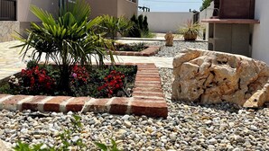 Ein Wohnhaus mit Terrasse, Stein-Barbecue und Garten mit roten Blüten und Grünpflanzen.