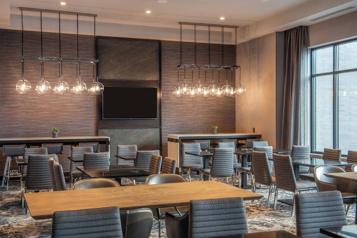 A modern hotel conference room with multiple tables and chairs, a large flat-screen TV, and a large window with curtains.