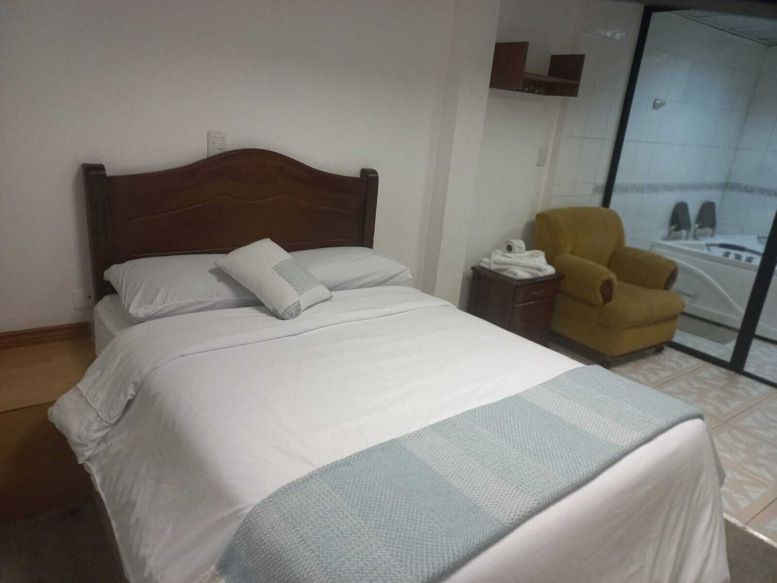 Una habitación de hotel con una cama, una silla y un baño visible a través de una puerta de vidrio.