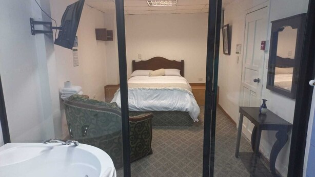 A hotel room with a bed, a small table, a mirror, a bathroom with a bathtub, and a patterned floor. - Hotel El Geranio (Quito)