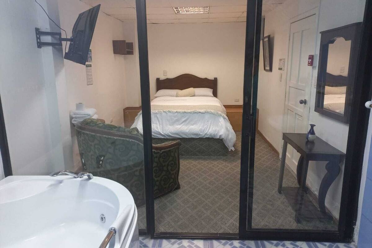 A hotel room with a bed, a small table, a mirror, a bathroom with a bathtub, and a patterned floor.