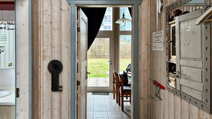 Kapea käytävä, jonka seinät ovat puuta ja jossa on laattalattia. K äytävän päässä on ovi, joka johtaa ulkotilaan, jossa on nurmikko ja pöytä.