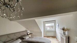 A bedroom with a large bed, a chandelier, a window, and a fireplace. - Living in Ohligs, attic apartment (Solingen)