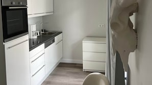 Une cuisine compacte avec des meubles blancs, un four noir et une machine à café. On y trouve une chaise blanche et une table avec un set de table.