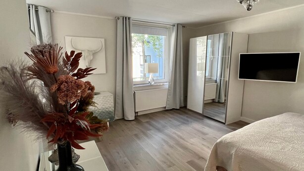 Une chambre à coucher avec un lit, un miroir, une télévision et une fenêtre avec des rideaux.