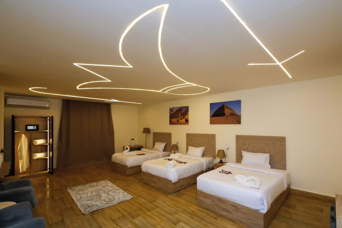 Habitación de hotel con tres camas, piso de madera, alfombra estampada y lámpara en el techo.