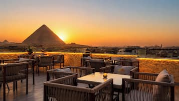 A rooftop terrace with wicker furniture, a stone wall, and a view of the pyramid at sunset.