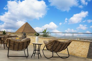 A rooftop terrace with wicker chairs, a small table, and a view of the pyramid.