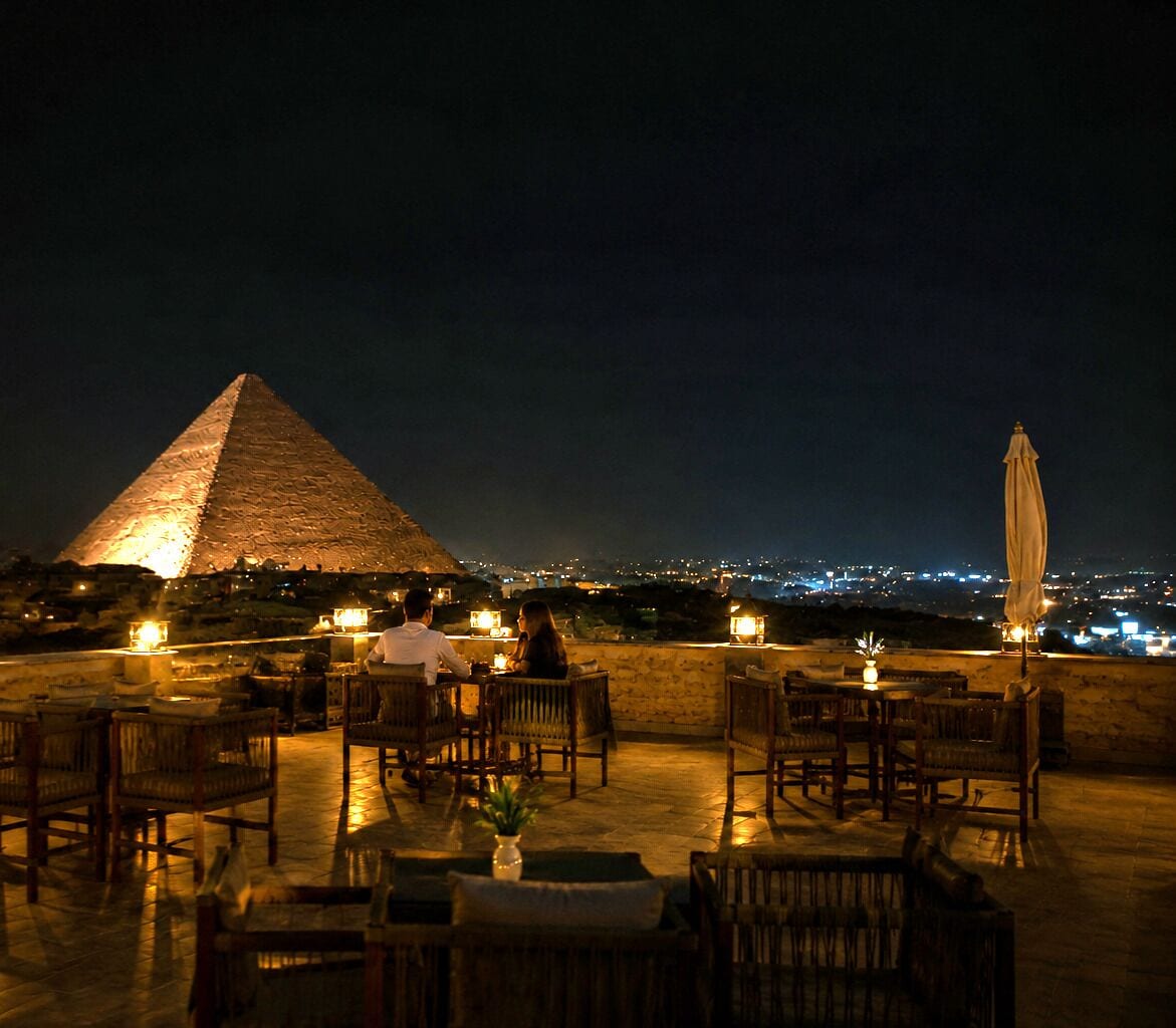 A dining area with tables and chairs, a pyramid in the background, and a cityscape view.