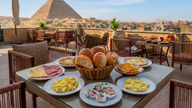 A dining table with various breakfast items and bread, set against a backdrop of a pyramid and cityscape. - Mazaya Pyramids View Inn (Giza)