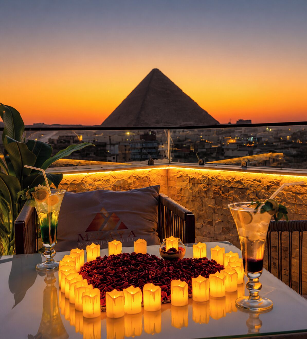 A table set with candles, drinks, and a heart-shaped arrangement of red roses, overlooking a cityscape and a pyramid at sunset.