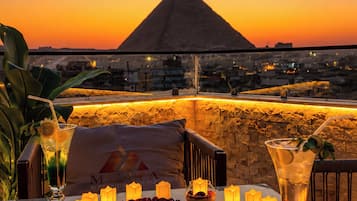 Una mesa dispuesta con velas, bebidas y un arreglo de rosas rojas en forma de corazón, con vista a un paisaje urbano y una pirámide al atardecer.