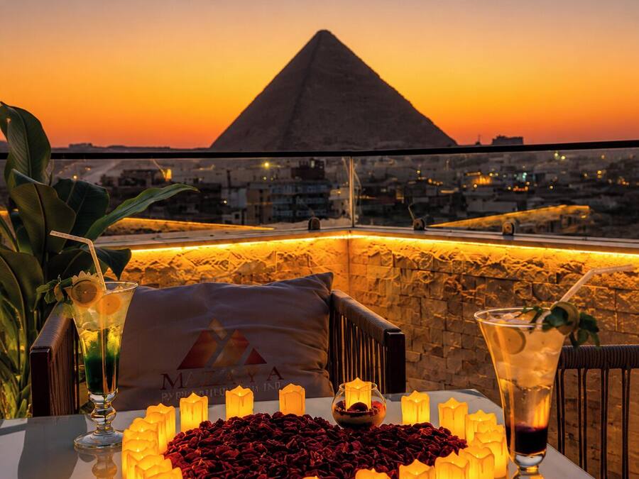 Una mesa dispuesta con velas, bebidas y un arreglo de rosas rojas en forma de corazón, con vista a un paisaje urbano y una pirámide al atardecer.