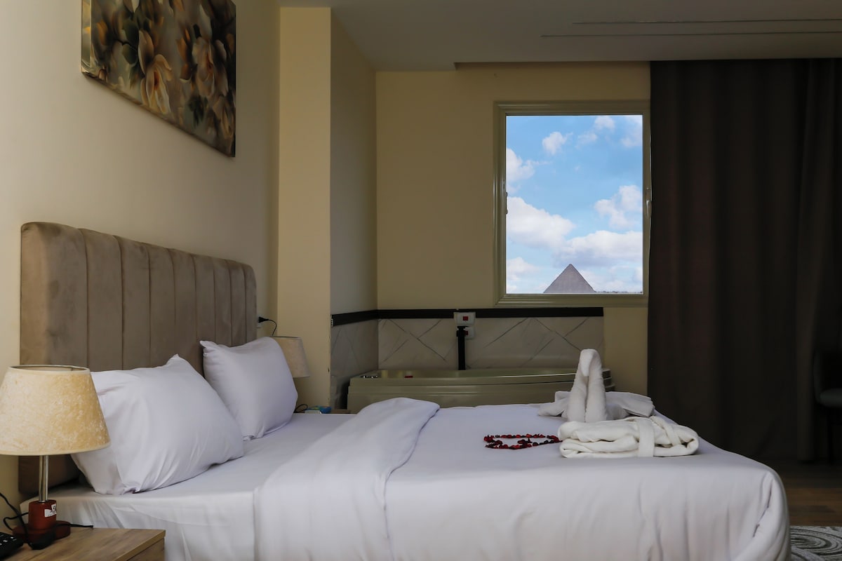 Ein Hotelzimmer mit Bett, Nachttisch, Lampe, Fenster mit Blick auf eine Pyramide und einem Bild an der Wand.