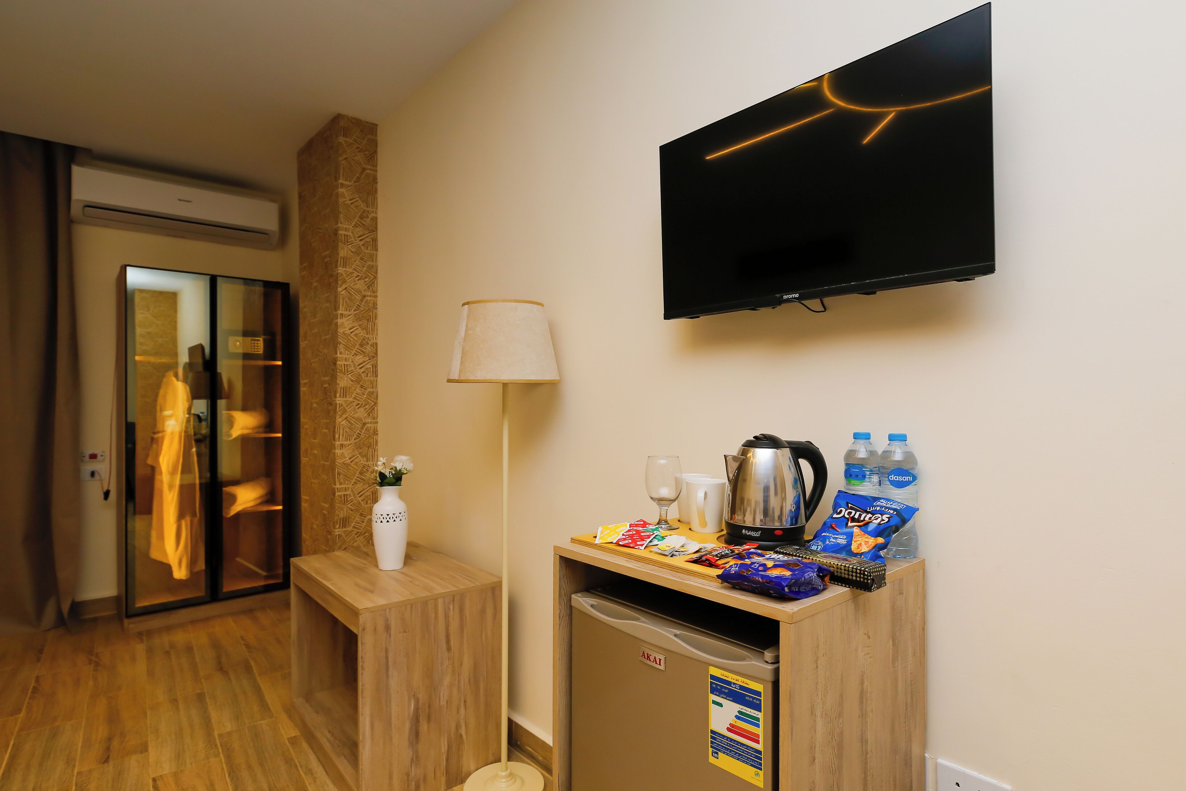 A hotel room with a minibar, a television mounted on the wall, a lamp, and a wooden floor.