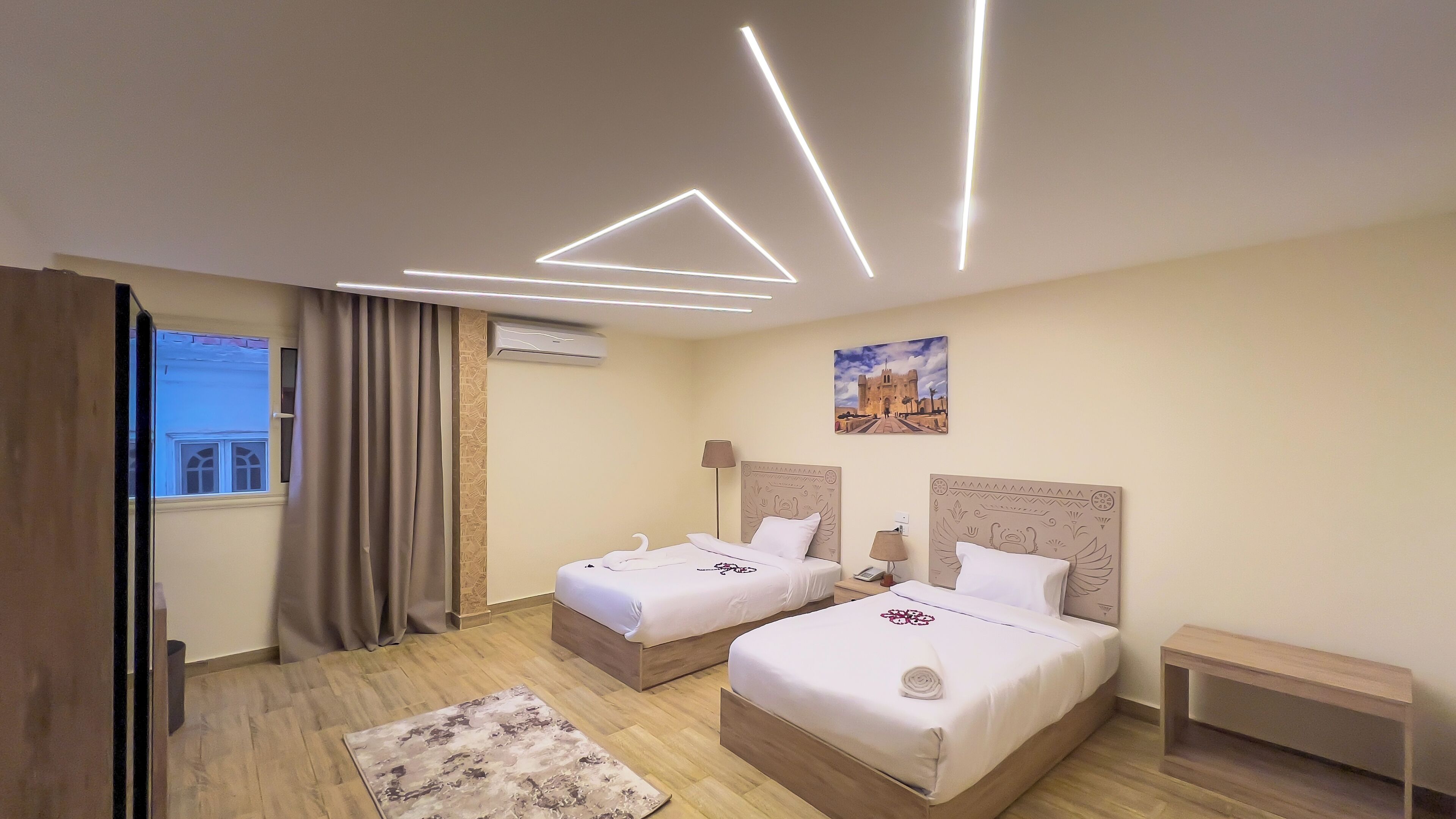 A hotel room with two beds, a wooden floor, a large window with curtains, a painting on the wall, and a modern ceiling light design.