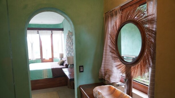 Een badkamer met een houten wastafelmeubel, een grote ovale spiegel en uitzicht op een slaapkamer door een boog.