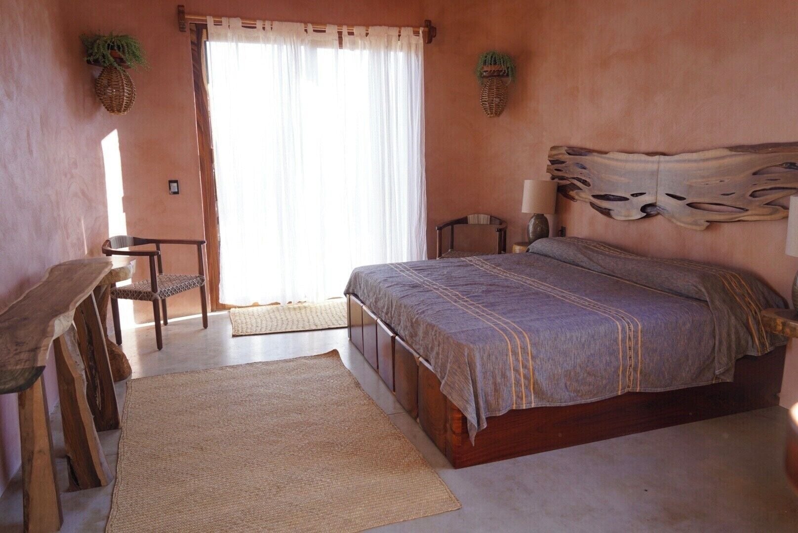 A bedroom with a bed, a desk, and a chair. There is a window with curtains, a wall-mounted lamp, and a woven rug on the floor.
