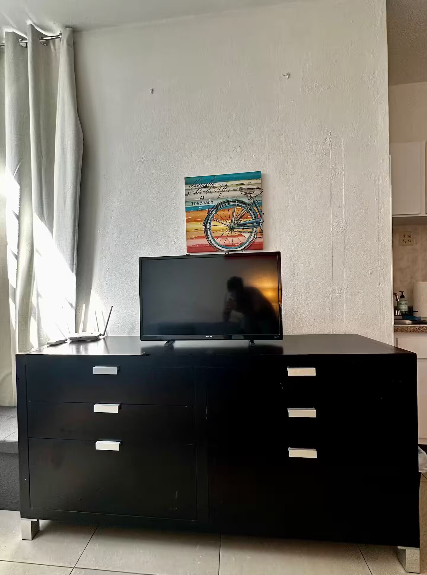A black TV stand with a flat-screen TV, a painting of a bicycle on a beach, and a white wall.