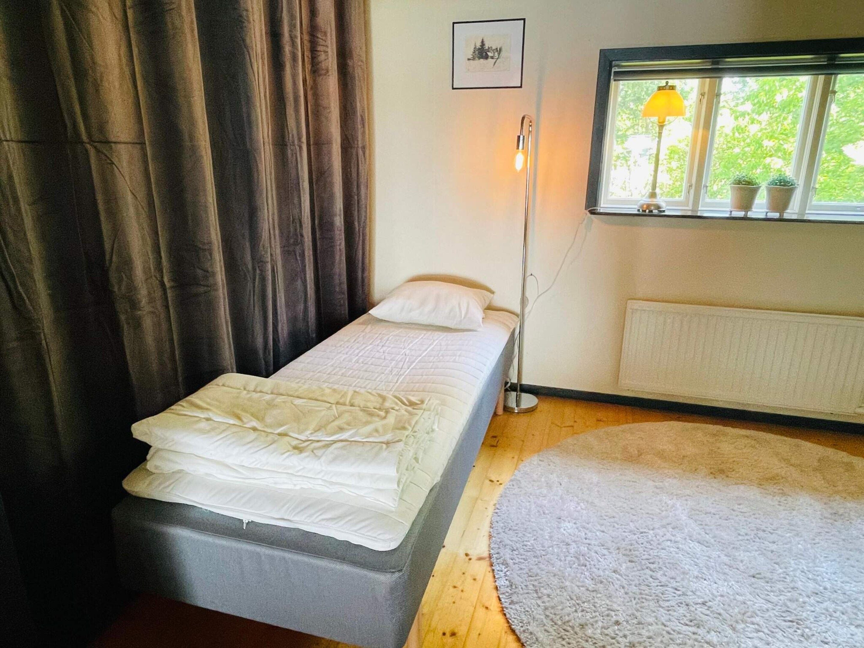 A small, single-bed room with a window, a radiator, and a floor lamp.