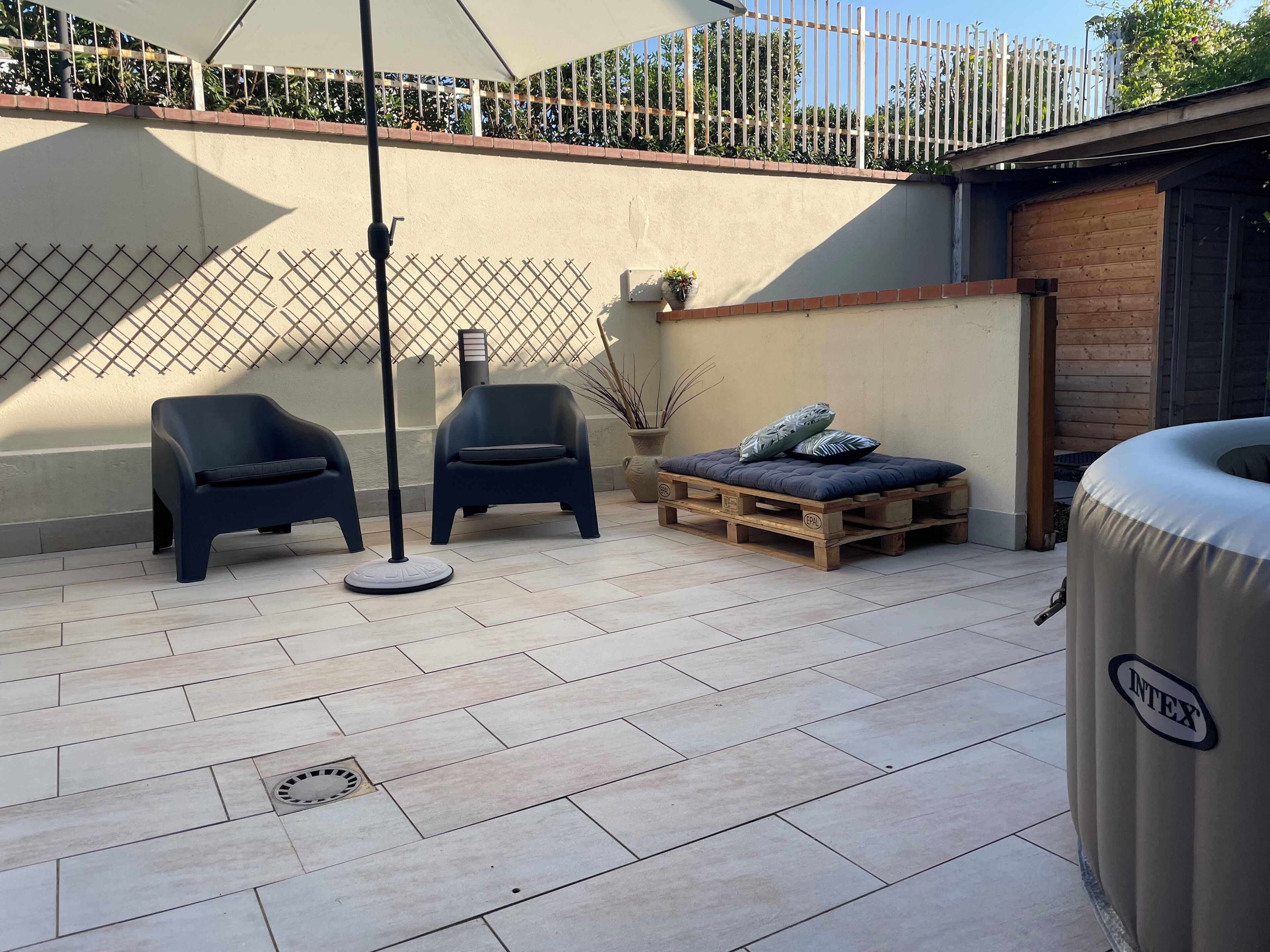 A patio area with two black chairs, a wooden pallet bed with pillows, a trash bin, and a fence in the background.