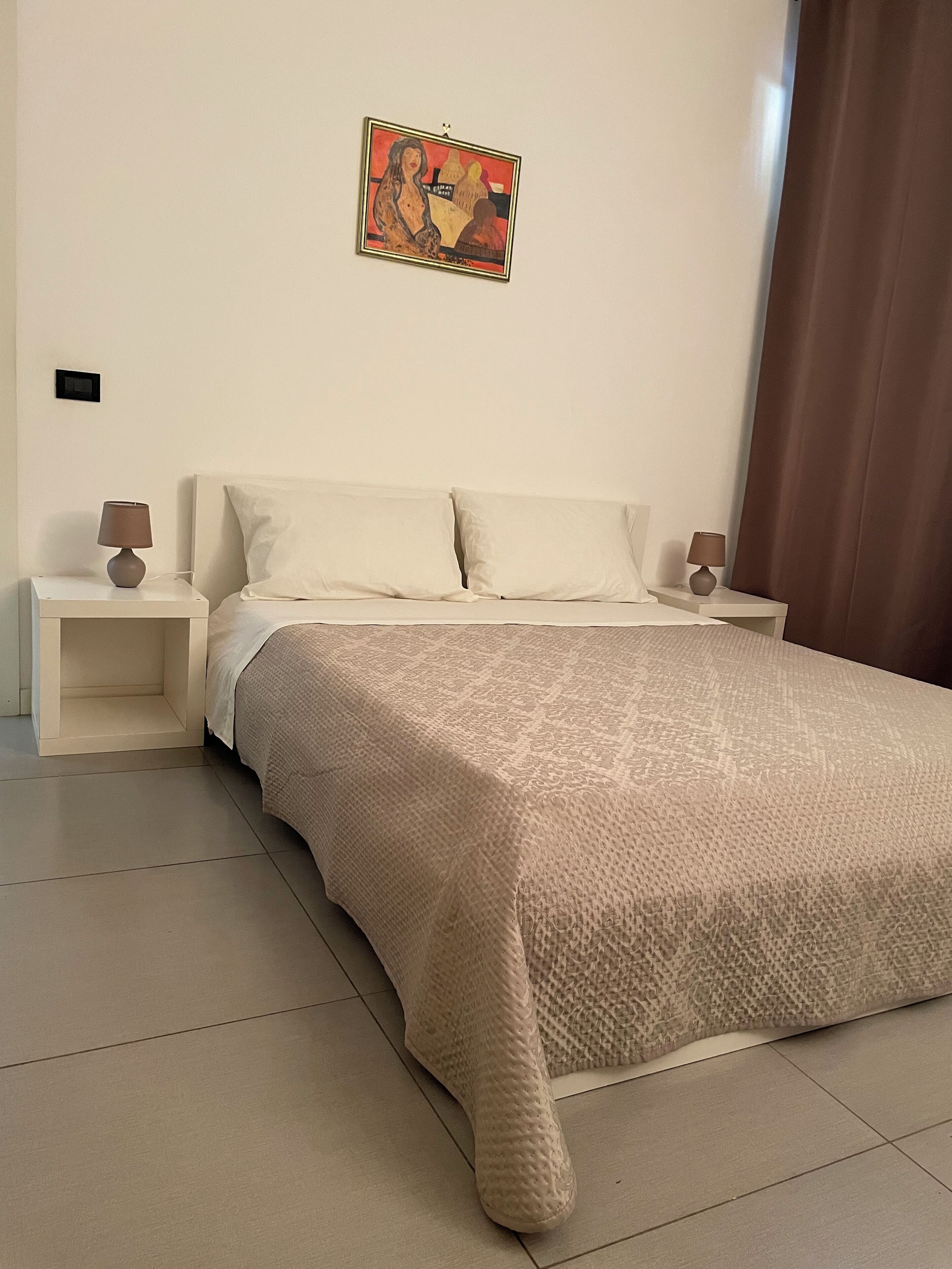 A bedroom with a large bed, two bedside tables with lamps, a wall-mounted painting, and a brown curtain.