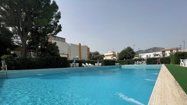 A swimming pool with clear blue water, surrounded by lounge chairs and umbrellas, with buildings in the background. - Cozy apartment just minutes from the beautiful Mondello beach. (Palermo)