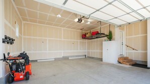 A spacious garage with a concrete floor, a red lawnmower, and a white door with a wooden staircase leading to a door. - Perfect Basecamp for Outdoor Fun (Driggs)