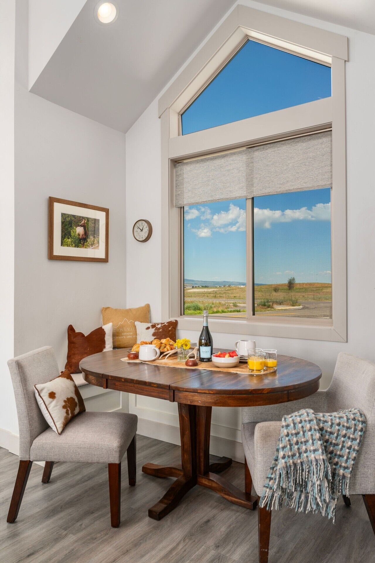 A dining area with a round table, chairs, a clock, a framed picture, and a view of a landscape through a window.