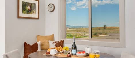 A dining area with a round table, chairs, a clock, a framed picture, and a view of a landscape through a window.