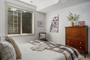 A bedroom with a large window, a bed with a quilt, a wooden cabinet, and a wall with a framed picture. - Perfect Basecamp for Outdoor Fun (Driggs)