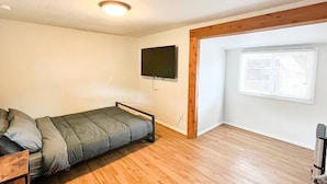 Une petite pièce, peu meublée, avec un plancher en bois, un lit simple, un téléviseur fixé au mur et une fenêtre avec des stores.