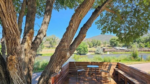 Une terrasse en bois, agrémentée de bancs et de tables, entourée de grands arbres et offrant une vue sur un lac et des collines.