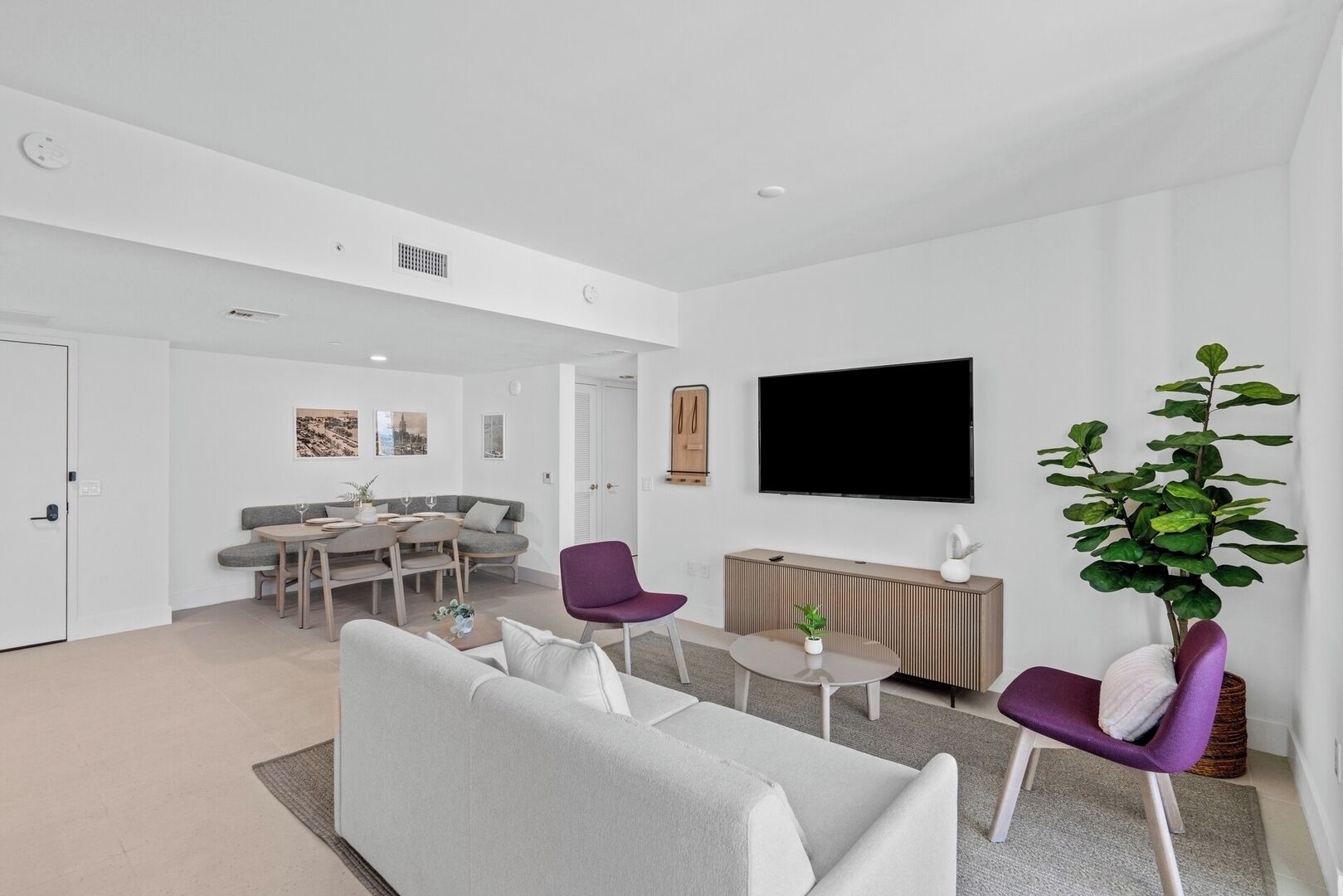 Un salon moderne avec un canapé, deux fauteuils violets, un coin repas comprenant une table et des chaises, un téléviseur à écran plat fixé au mur et une plante en pot.
