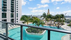 Ein Pool auf dem Dach mit klarem, blauem Wasser, umgeben von Palmen und Liegestühlen. Im Hintergrund sieht man einen hohen Turm und ein modernes Gebäude.