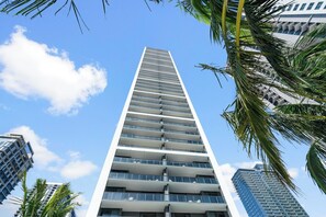 A modern high-rise building with multiple balconies, surrounded by palm trees and other high-rise buildings.