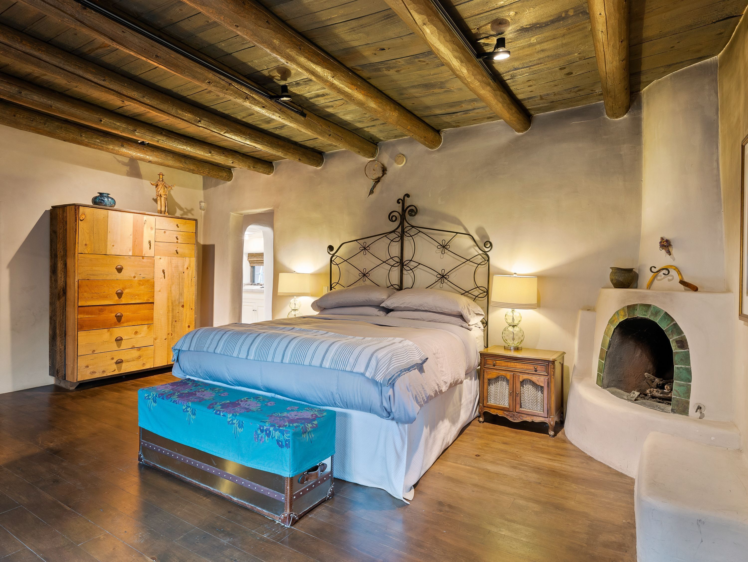 Ein Schlafzimmer mit Holzboden, einem Bett mit blau-weißer Bettdecke, einer hölzernen Kommode, einem kleinen Tisch mit Lampe und einem Kamin mit grün gefliestem Bogen.