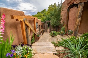 Un sentier en bois mène à un bâtiment rustique doté d’un porche en bois et d’un jardin où poussent diverses plantes et fleurs.