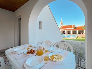 A breakfast table set with plates, croissants, fruit, and juice, overlooking a swimming pool and a building with a tower. - Rose Sun Retreat - apartment with shared pool (Viana do Castelo)