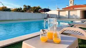 Une zone avec une piscine, des chaises longues, une table avec deux verres de jus d’orange et, en arrière-plan, une église dotée d’un clocher.