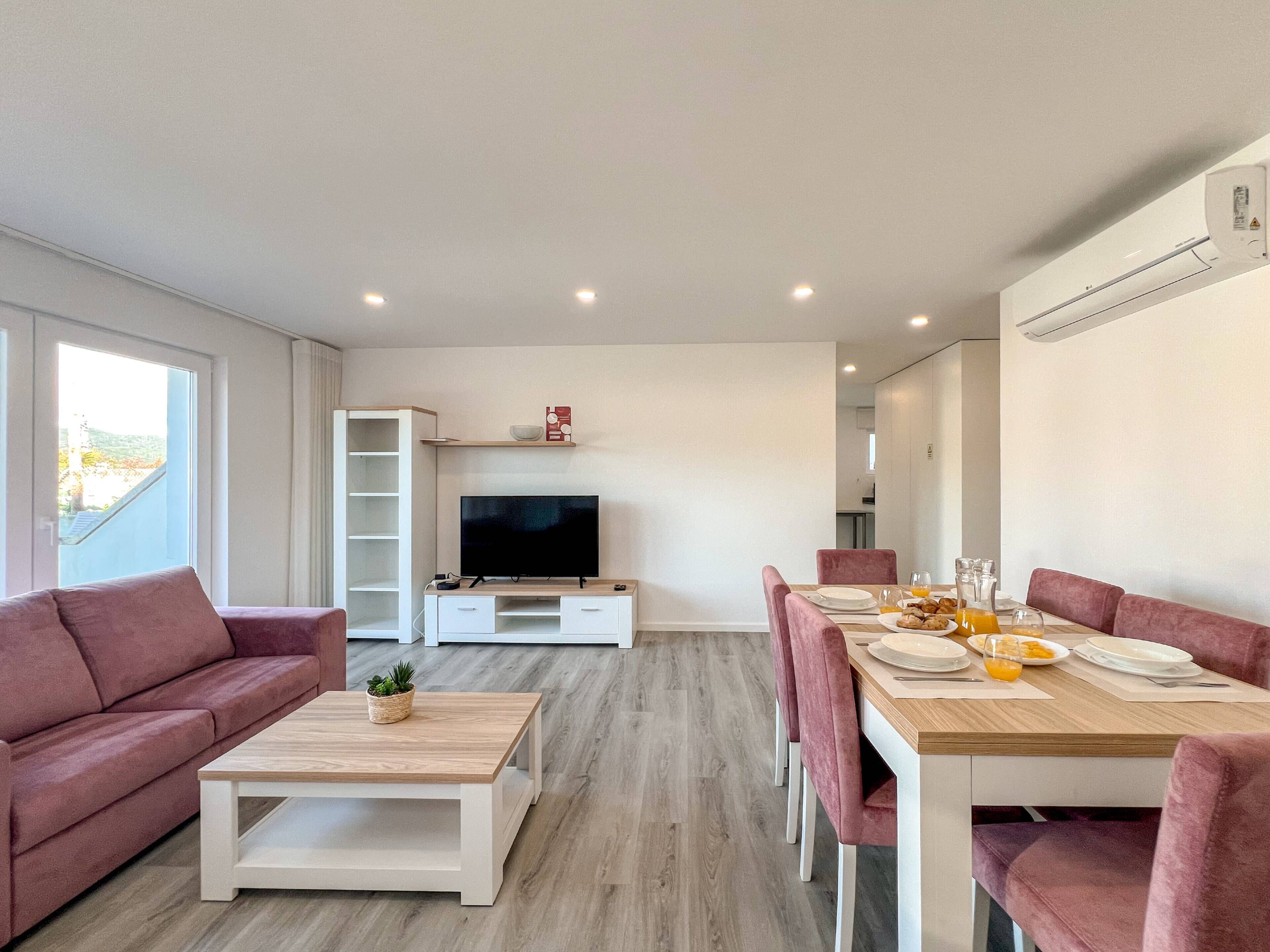 A modern living room with a TV, sofa, coffee table, and dining area with pink chairs and a wooden table.