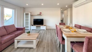 Un salon moderne comprenant un téléviseur, un canapé, une table basse et un coin repas avec des chaises roses et une table en bois.