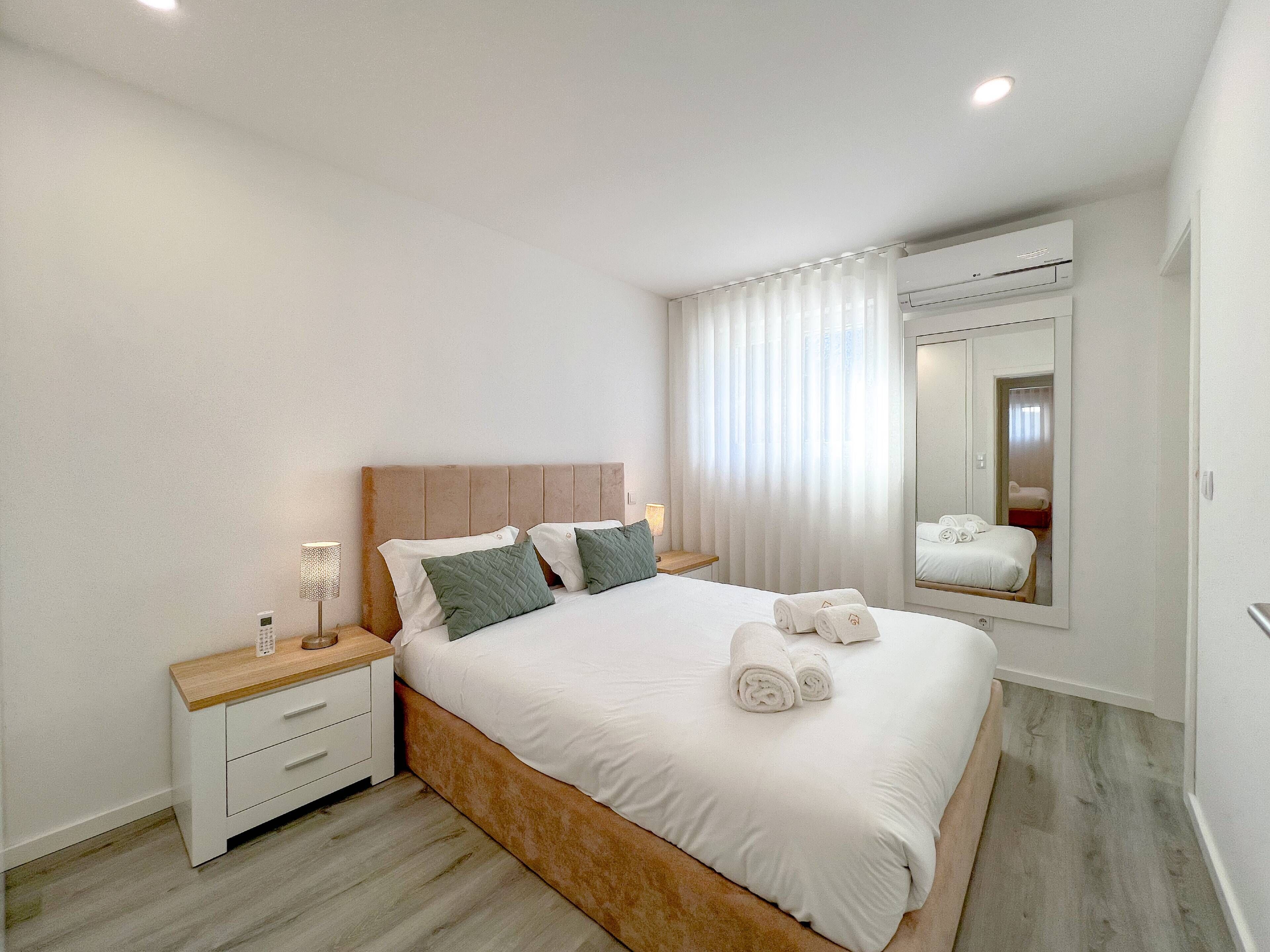 Ein modernes Schlafzimmer mit einem Holz-Kopfteil, einem großen Bett mit weißer Bettwäsche, einem Nachttisch mit Lampe und einem Spiegel an der Wand.